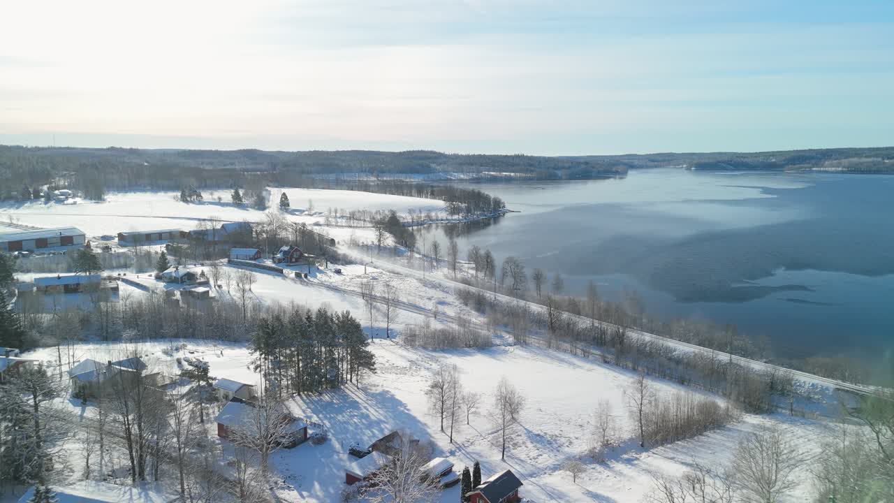 Un drone sorvola le case di legno innevate del villaggio di Liane mentre un treno passeggeri viaggia lungo il lago ghiacciato Teåkersjön nella zona rurale di Dalskog Mellerud in Svezia durante una giornata invernale - vista aerea ad alta angolazione