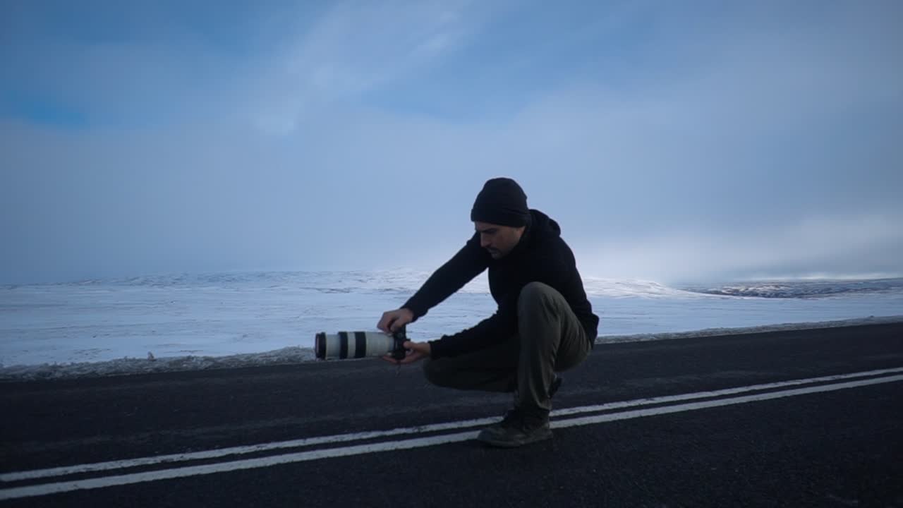 A low-angle shot captures a photographer crouched by the side of a desolate road in North Iceland, seemingly adjusting their camera or framing a shot