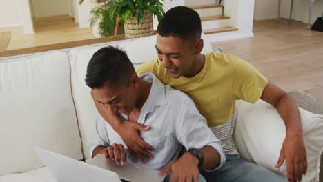 Smiling-mixed-race-gay-male-couple-sitting-on-sofa-using-laptop-and-laughing