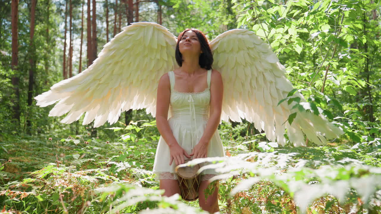 Angel woman with white wings in sunlit forest examines talking drum, lifts rope, positions instrument between legs, then shifts focus to kalimba, preparing gentle woodland performance