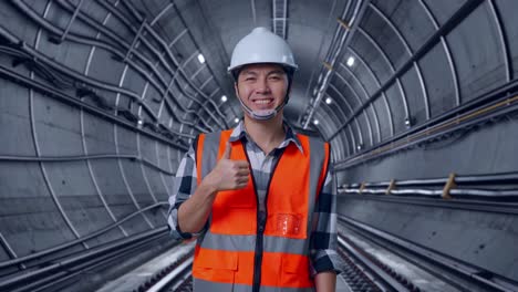 asian male engineer with safety helmet smiling and showing thumbs up gesture to the camera while standing in underground subway tunnel