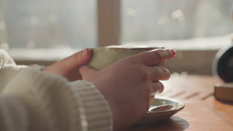 close up of person gently holding tea cup with both hands as warm morning sunlight shines through window creating soft glow on table surface and ceramic