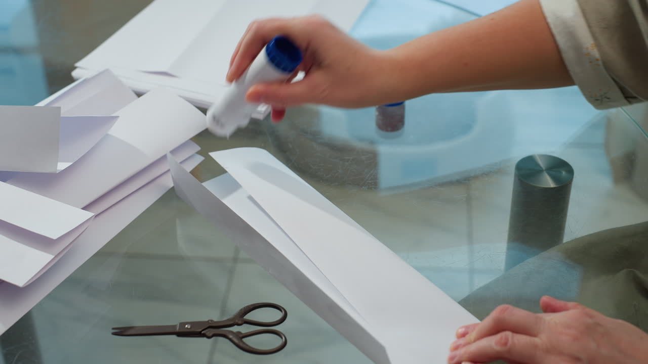 Hand view of aged woman applying glue on paper craft on table, scissors and white paper scattered on surface, showcasing a crafting session in a peaceful home setting with focus on hands