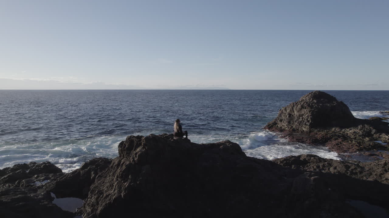fantastica toma aerea sobre la costa de galdar y donde se puede ver a una mujer sentada en una gran roca y admirando las olas del mar y el paisaje