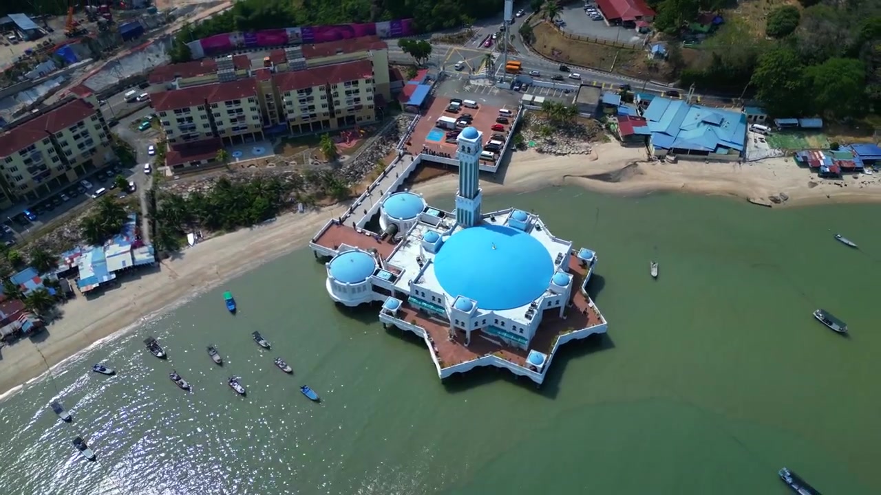 Den flydende moské i Tanjung Bungah, Penang, Malaysia, med traditionelle fiskerbåde langs kysten og moderne bygninger i baggrunden. Speed ramp time-lapse. Smuk cirkulær droneoptagelse fra luften.