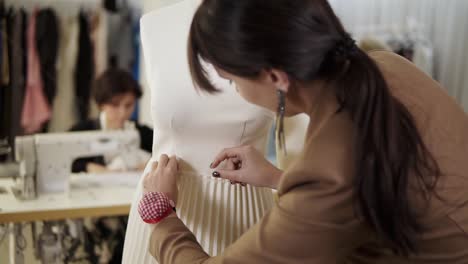 Creative-fashion-designers-are-working-together-in-the-tailor-studio.-Long-haired-woman-pinning-on-the-white-skirt-on-mannequin-using-pins-on-her-hand-pillow-on-wrist.-Woman-working-on-sewing-machine-on-a-blurred-background