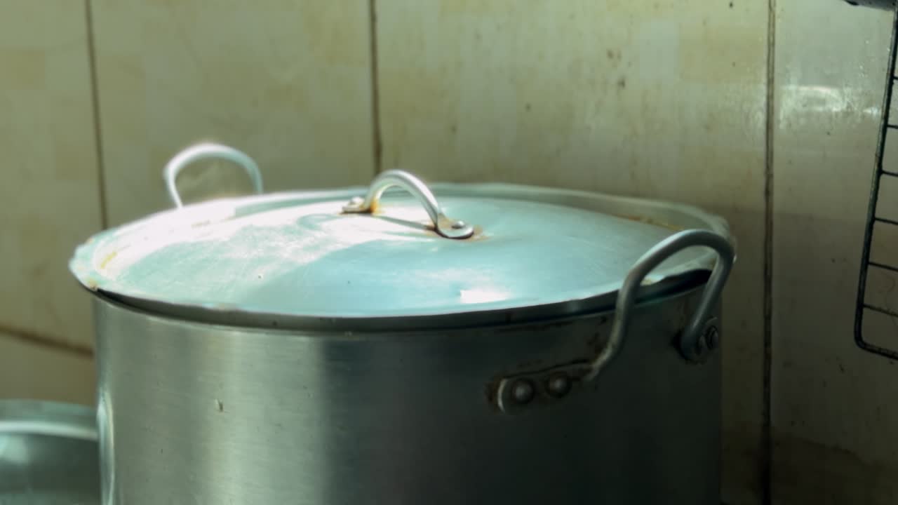 Primo piano di una pentola da cucina in alluminio con coperchio appoggiata a una parete piastrellata della cucina, scattato in interni con luce soffusa, texture di metallo usurato e un'atmosfera pratica di cucina casalinga.