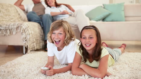 siblings lying on the floor and watching tv with parents behind them