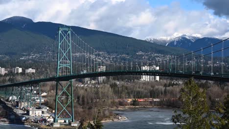 timelapse rooling waves agains the rocks under the blue pillar of the lions gate suspension bridge while traffic are driving over and clouds running over the with snow covered rocky mountain