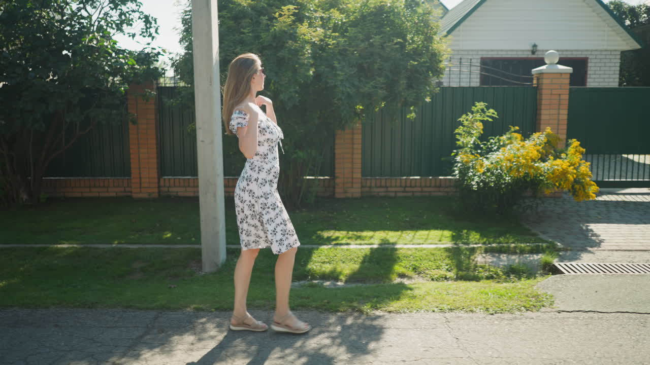 Side view of stylish woman walking under sunlight while adjusting her floral dress, holding and releasing cloth tie, wearing sunshades, passing green yard and brick wall of charming neighborhood home