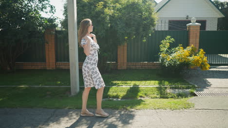 side view of stylish woman walking under sunlight while adjusting her floral dress, holding and releasing cloth tie, wearing sunshades, passing green yard and brick wall of charming neighborhood home