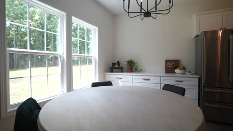 orbiting shot of dining room table in the kitchen of the house, with bright windows showing the view of the trees in the backyard and patio