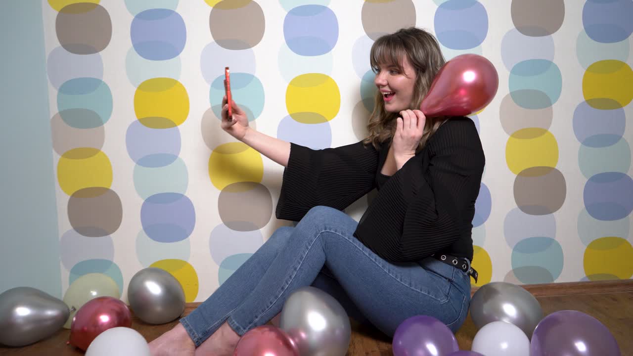Giovane donna seduta tra palloncini e con un telefono in mano, su sfondo colorato.