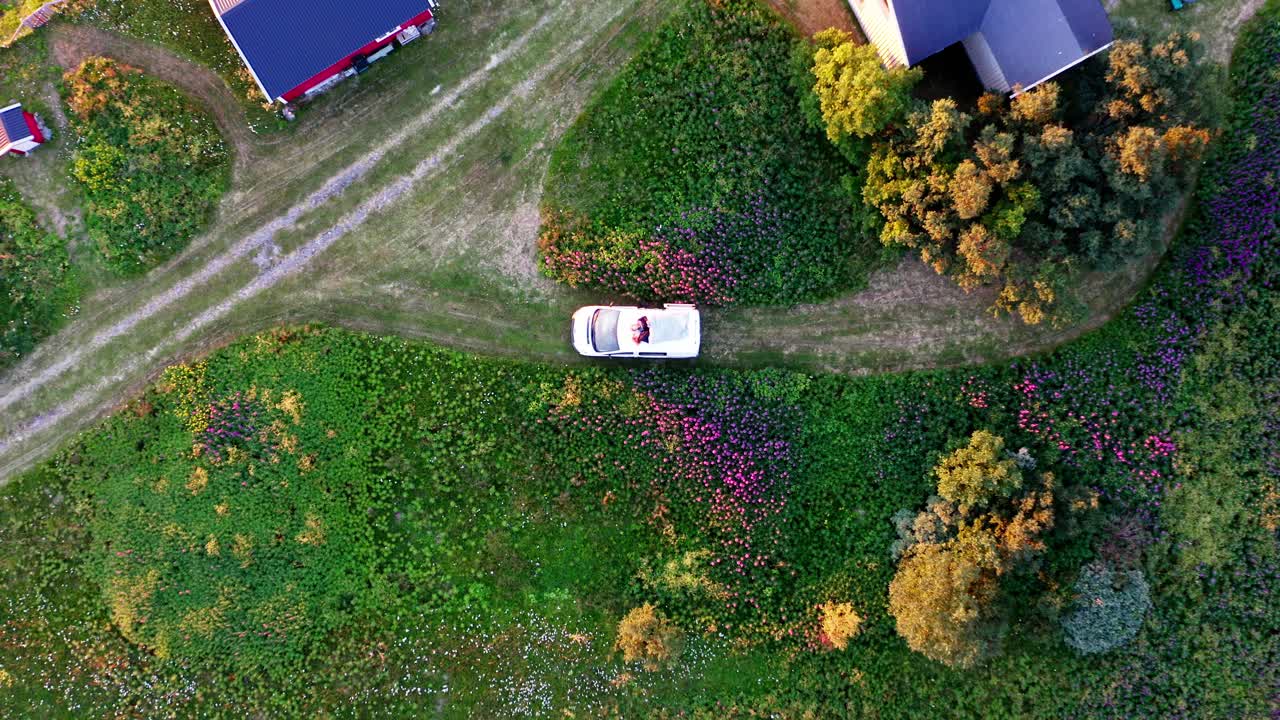 Romantic Couple Lying And Relaxing On Roof Of White Car Outside Their House Surrounded By Colorful Flowers On A Cold Sunny Day - top-down drone pullback (ascend)