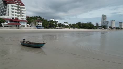a small fishing boat rests on a quiet beach with modern buildings and resorts in the background under a cloudy sky.