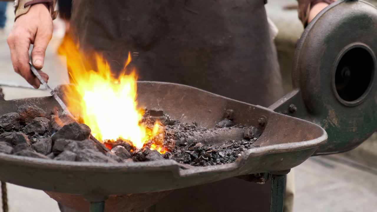Slow-motion static close-up as a blacksmith turns the hand crank of a coal forge, fanning the fire; bright flames swell while coals glow and light smoke drifts