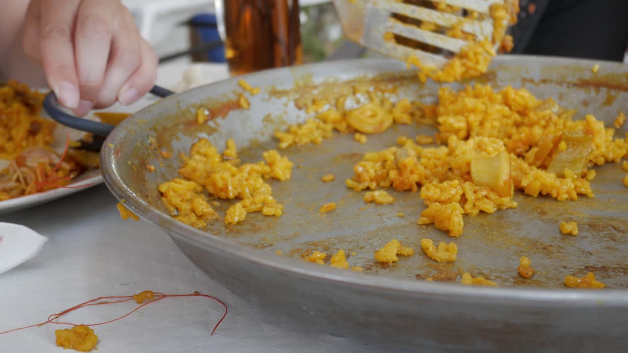 A person is eating paella from a large pan, with only a small portion of rice remaining