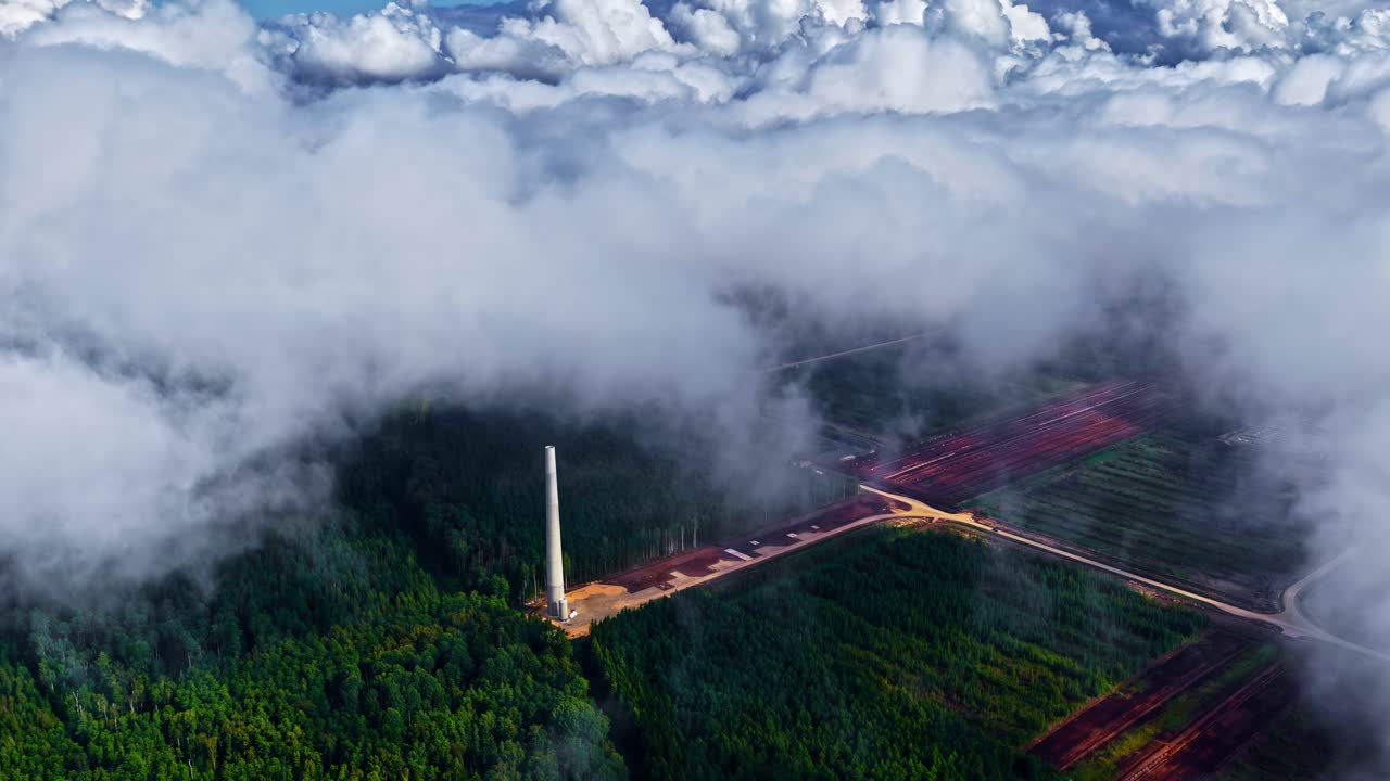 Dramatic aerial view above a dense forest partially covered in thick, billowing clouds. A tall, slender white tower or chimney, standing out sharply against the natural surroundings