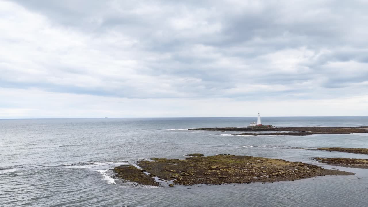 Drone camera glides over rocky shoreline toward a distant lighthouse under overcast skies, capturing expansive seascape and tranquil coastal environment in natural daylight