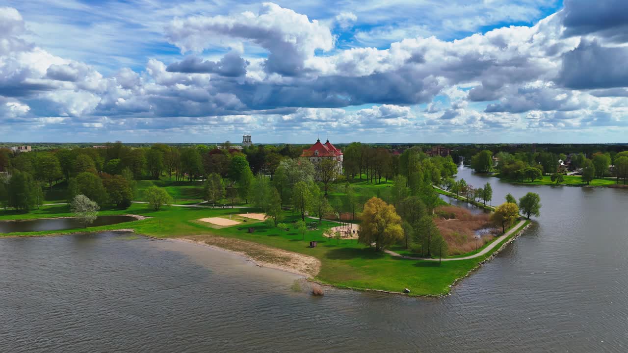 wide-angle panorama captures Biržai's Wibyholms Manor in spring, with vibrant green lawns, a red-roofed estate, and a tranquil lake under a dynamic sky filled with white and grey clouds