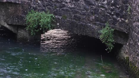 Flacher,-Kristallklarer-Fluss,-Der-Unter-Dem-Alten-Steinbrückentorbogen-Mit-Pflanzen-In-Der-Englischen-Ländlichen-Landschaft-Fließt