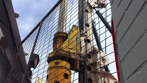 looking through a fence at a hydraulic rotary drilling rig as it rotates borer column and raises a drill bit filled with dirt