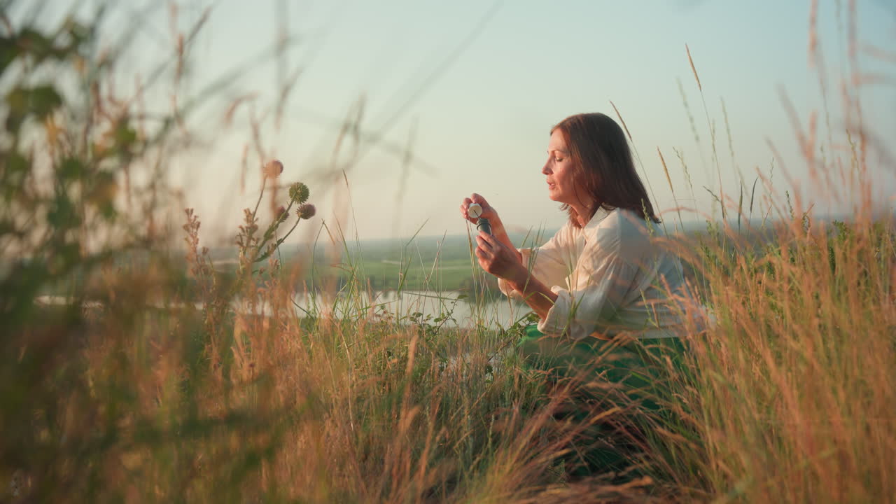 Sunset lit lady in green skirt squatting river bank opening bubble container then blowing soap bubbles drifting over blurred valley under warm golden light among wild grass and flowers