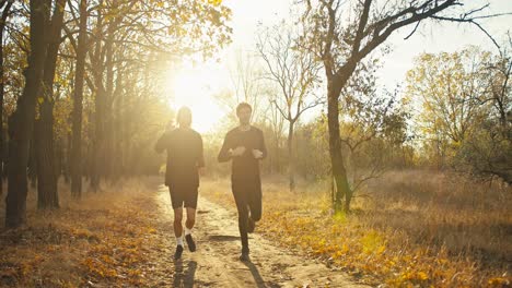 Two-guy-friends-in-black-sportswear-are-running-to-their-destination-during-their-morning-jogging-in-an-autumn-park-with-fallen-brown-leaves-at-Sunrise-in-autumn