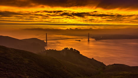 5k time lapse shot of golden gate bridge during beautiful yellow sunrise and boat cruising below bridge - epic lighting of sky with flying clouds - aerial wide shot