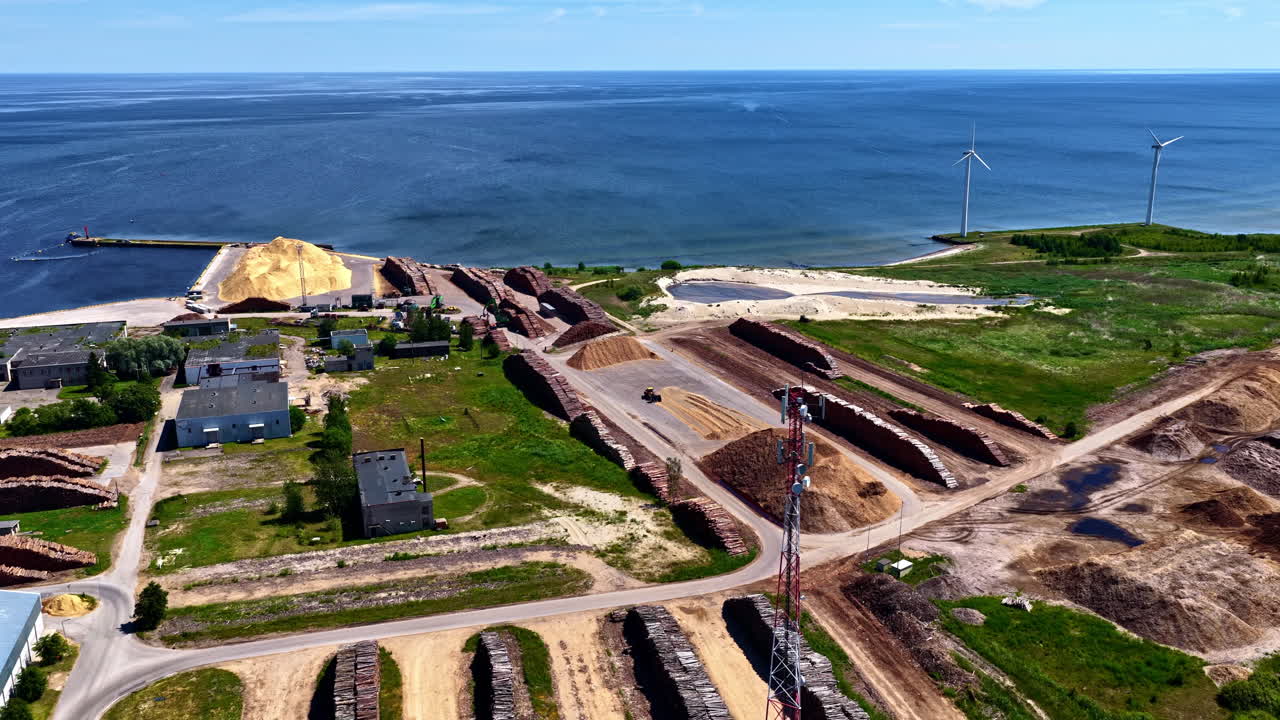Waterfront construction area with scattered cut logs and two windmills in the background