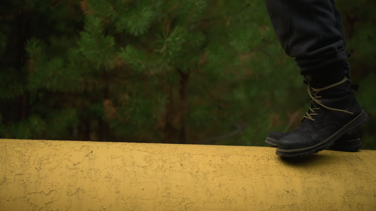 boots striding on yellow pipeline forest, lateral closeup of black boots and yellowlaced soles moving with steady cadence across mossdotted pipe, gentle green backdrop and soft bokeh evoke