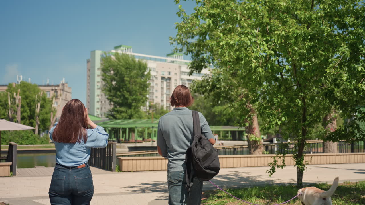 coppia passeggia lungo il fiume, coppia urbana si gode una passeggiata sul lungomare, coppia passeggia accanto al fiume con la città sullo sfondo, due persone fanno una passeggiata lungo il fiume con vista sulla città e sugli alberi