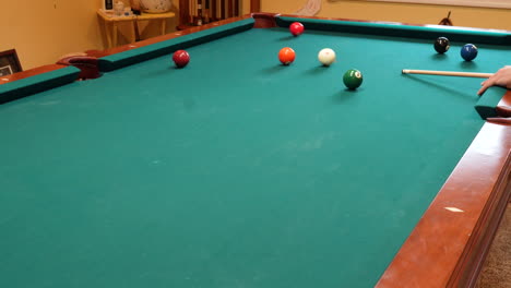 man playing 8 ball pool shoots several solid balls into pockets on a brunswick table for billiards with green felt in basement of home, open bridge hand and wooden cue stick, wide angle