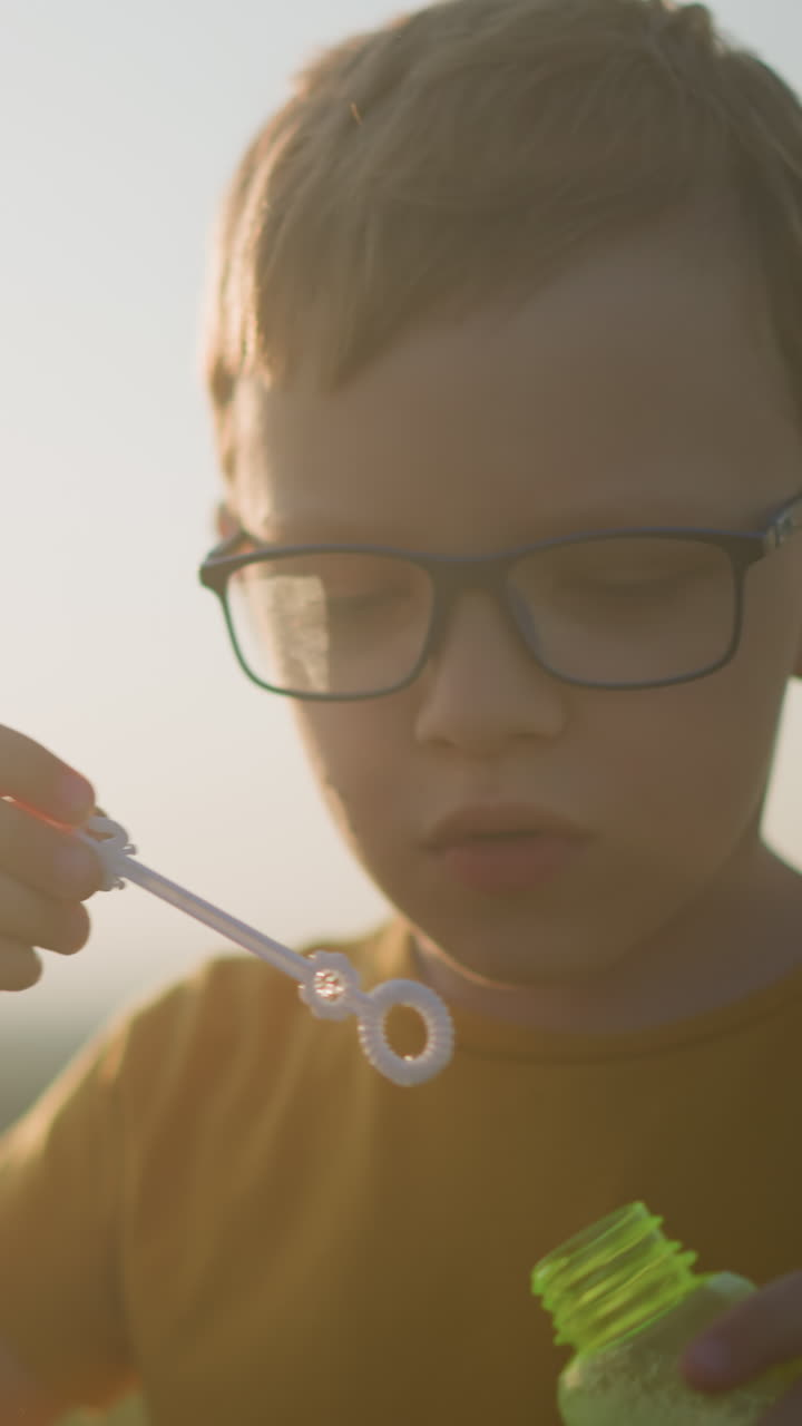 A boy in a yellow shirt and glasses carefully dips a wand into a foaming bottle, preparing to blow bubbles into the air. The image captures a serene moment of concentration and joy during a sunset