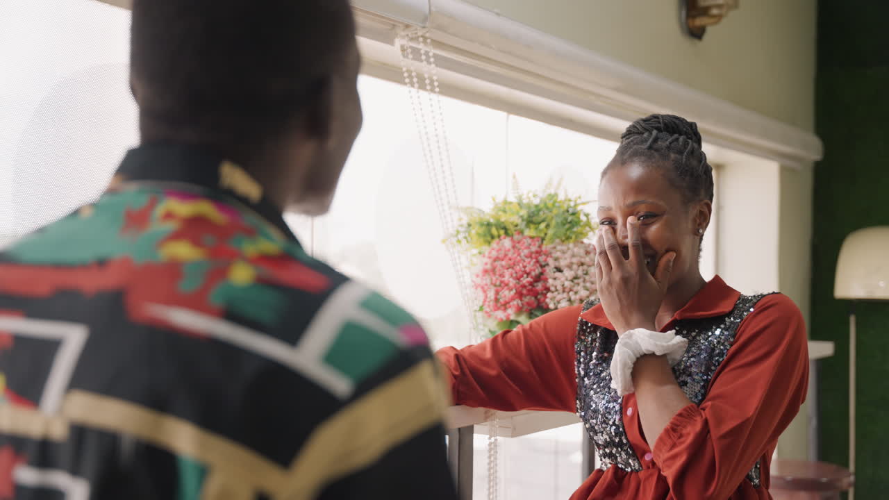 Mujer afroamericana sonriendo junto a la ventana, tono juguetón mientras se apoya en el marco y conversa con un hombre con camisa estampada, risa alegre, mano cubriendo la boca ante un cumplido, luz solar cálida, energía de cita afectuosa