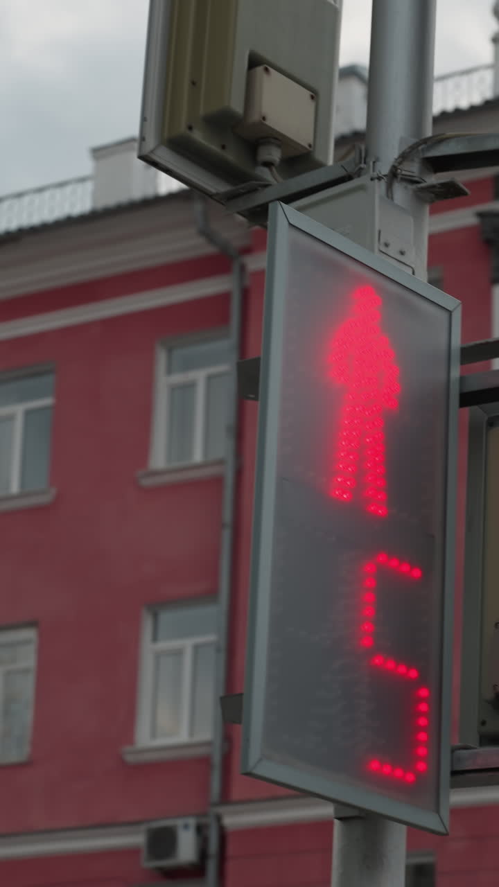 vista cercana de un poste de señal de cruce de peatones con un borde amarillo de neón y un semáforo digital que muestra una cuenta atrás. un fondo de edificio de ciudad de color rojo bajo un cielo nublado
