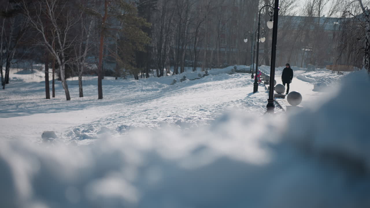 blurred foreground of snowdrifts in park path with child in colorful snowsuit playing while adult watches near lampposts and soft winter daylight casting gentle haze over serene seasonal landscape
