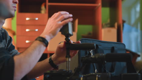 studio guy reaching into camera equipment bag on table while adjusting camera gear. focus on hand and equipment. background features green and orange walls with soft indoor lighting