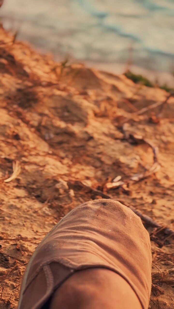 Un primo piano di un piede che indossa una scarpa slip-on beige che poggia su terra secca cosparsa di ramoscelli e vegetazione rada sotto una luce calda e soffusa