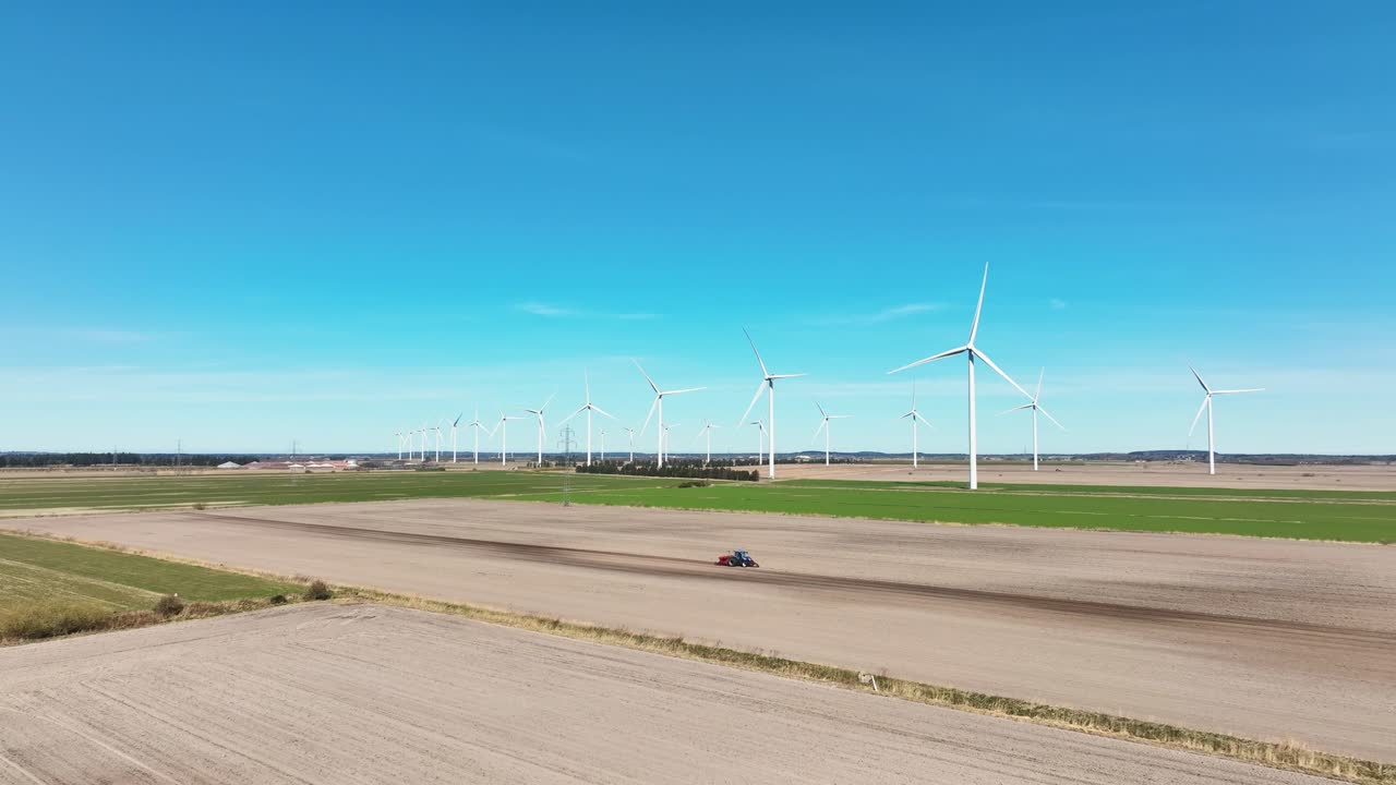 A drone view captures a farmer's tractor working next to a large wind farm in springtime, with the vibrant fields and towering turbines creating a striking contrast.