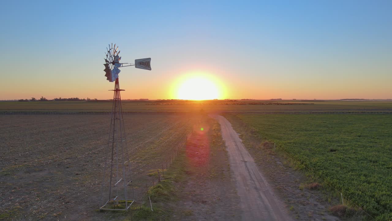 이른 아침 일출 동안 수정된 잔디밭 한가운데에서 바람과 함께 회전하는 소박한 물 펌핑 풍차를 캡처하는 공중 팬 샷