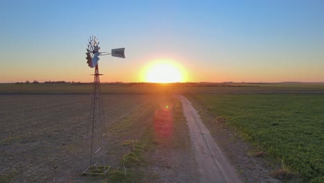 tiro panorámico aéreo que captura el molino de viento de bombeo de agua rústica girando con el viento en medio del campo de hierba fertilizado durante el amanecer temprano en la mañana