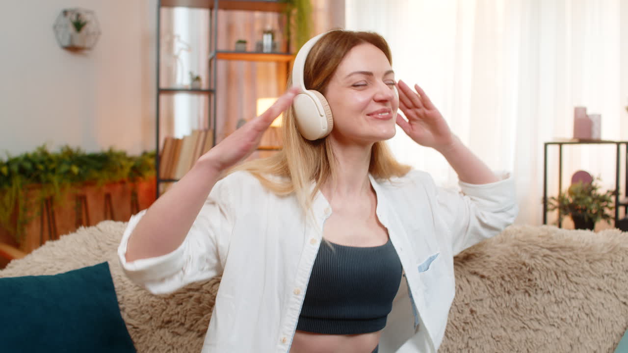 Adult woman dancing on home sofa with wireless headphones enjoying favorite song energetic mood