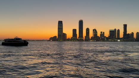 A-boat-sails-peacefully-along-the-Hudson-River,-while-the-silhouette-of-Jersey-City-glows-in-the-background