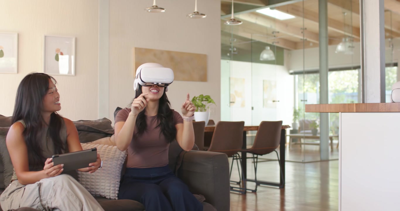 Amici di diverse etnie a casa che guardano una donna asiatica con un visore VR bianco, che indica, afferra e dimostra.