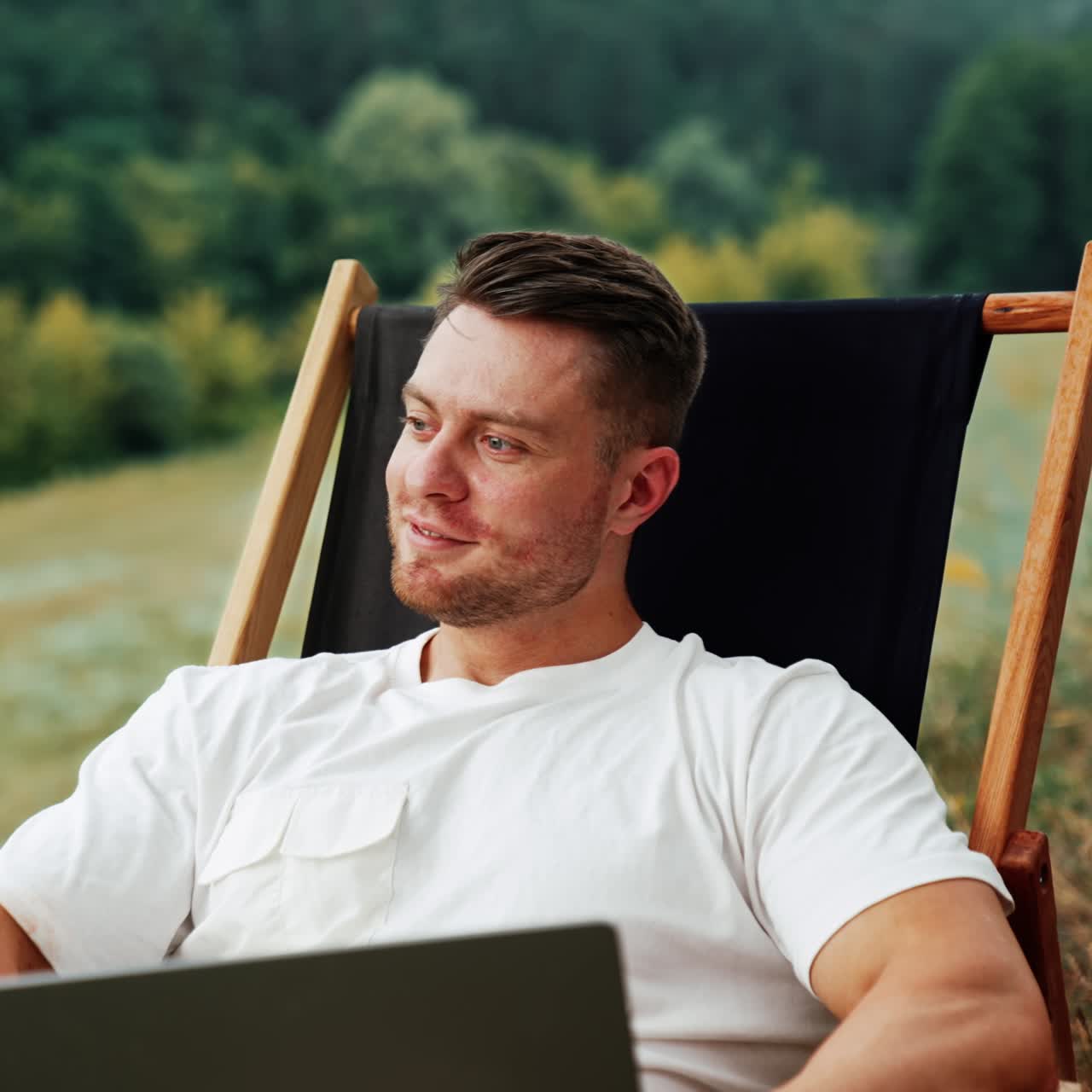 Thoughtful Caucasian man sits holding a laptop smiling dreamily. Freelancer working remote in a comfortable natural surroundings