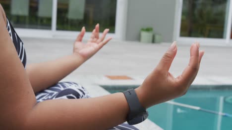 african american plus size woman practicing yoga, sitting meditating next to swimming pool in garden