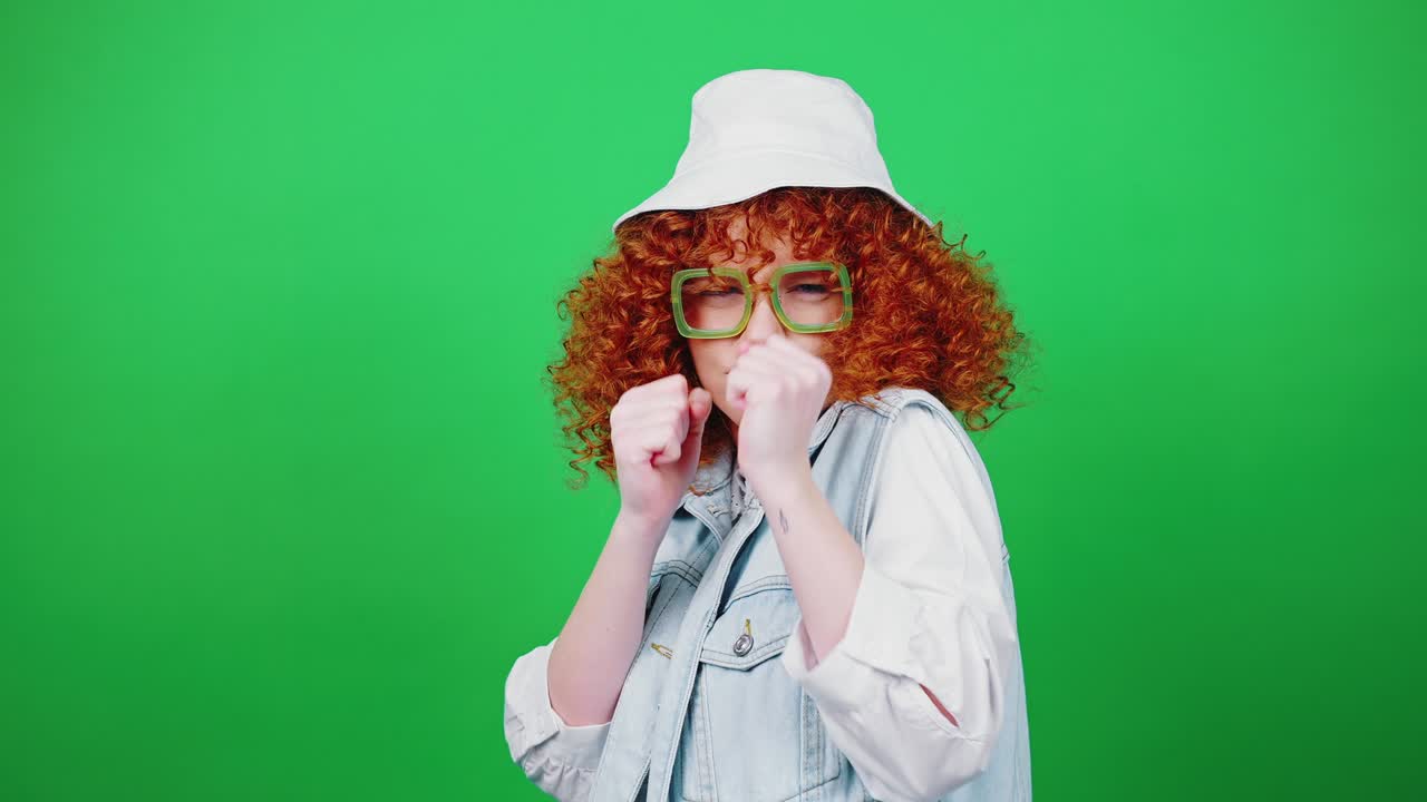 juguetona mujer pelirroja rizada con gafas y sombrero de panamá bromeando, boxeando ante la cámara y riendo, fondo de clave de croma verde