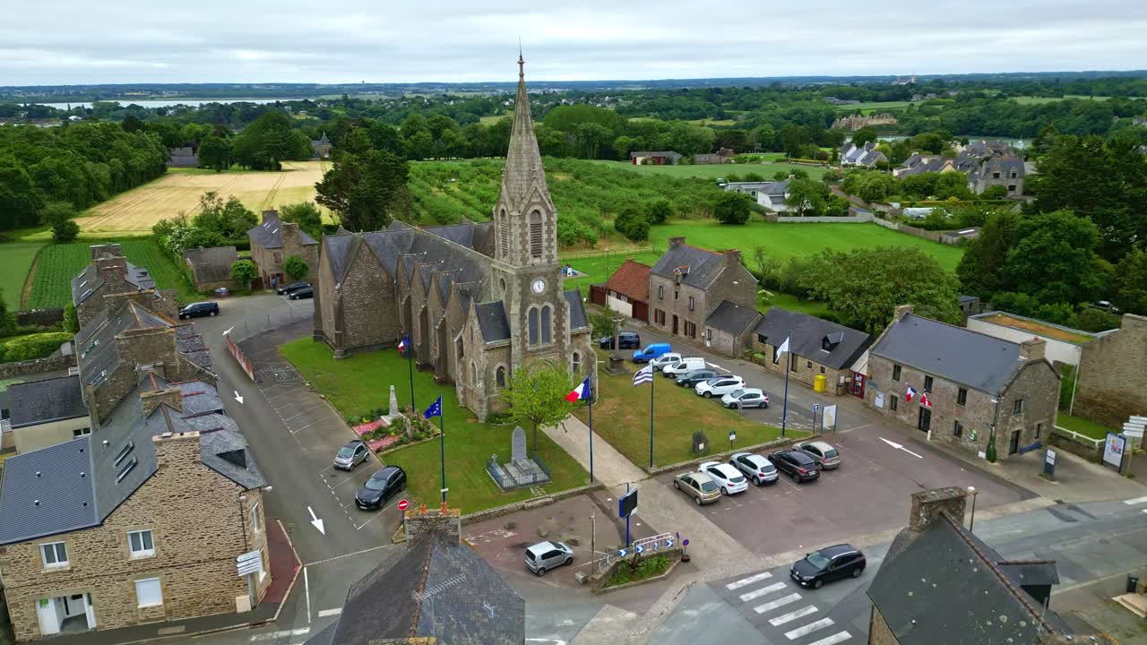 Drone che avanza con una leggera angolazione verso il basso verso la chiesa e la piazza di Notre-Dame-du-Guildo a Saint-Cast-le-Guildo, con bandiere e campi circostanti. Bretagna, Francia.