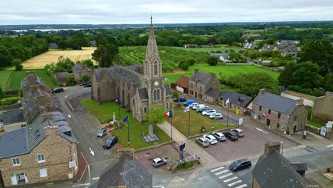 drone advances in slight downward angle toward notre-dame-du-guildo church and square in saint-cast-le-guildo, with flags and surrounding fields. brittany in france